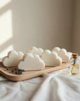 Cloud-shaped white baby soap arranged on a light wooden tray with dried lavender sprigs, next to a small glass bottle with oil, set on white fabric under soft natural light.