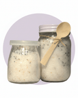 Two clear glass jars filled with white salt-like crystals, one small jar with a translucent lid and one larger jar with a white lid and tied wooden spoon, set against a light lavender circular backdrop on a white background.