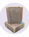 Two rectangular handmade soap bars stacked on top of each other, featuring a marbled swirl pattern of brown, muted green, and blue-gray, set against a light lavender circular background on a white canvas.