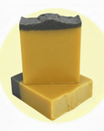 Two rectangular bars of handmade soap stacked on a pale yellow circular background. The top bar is upright, showing a dark charcoal gray top layer and a warm golden-yellow body.