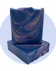Two rectangular handmade soap bars stacked on top of each other, featuring a deep blue and purple marbled swirl pattern with embedded gold glitter, set against a light blue circular graphic on a white background.
