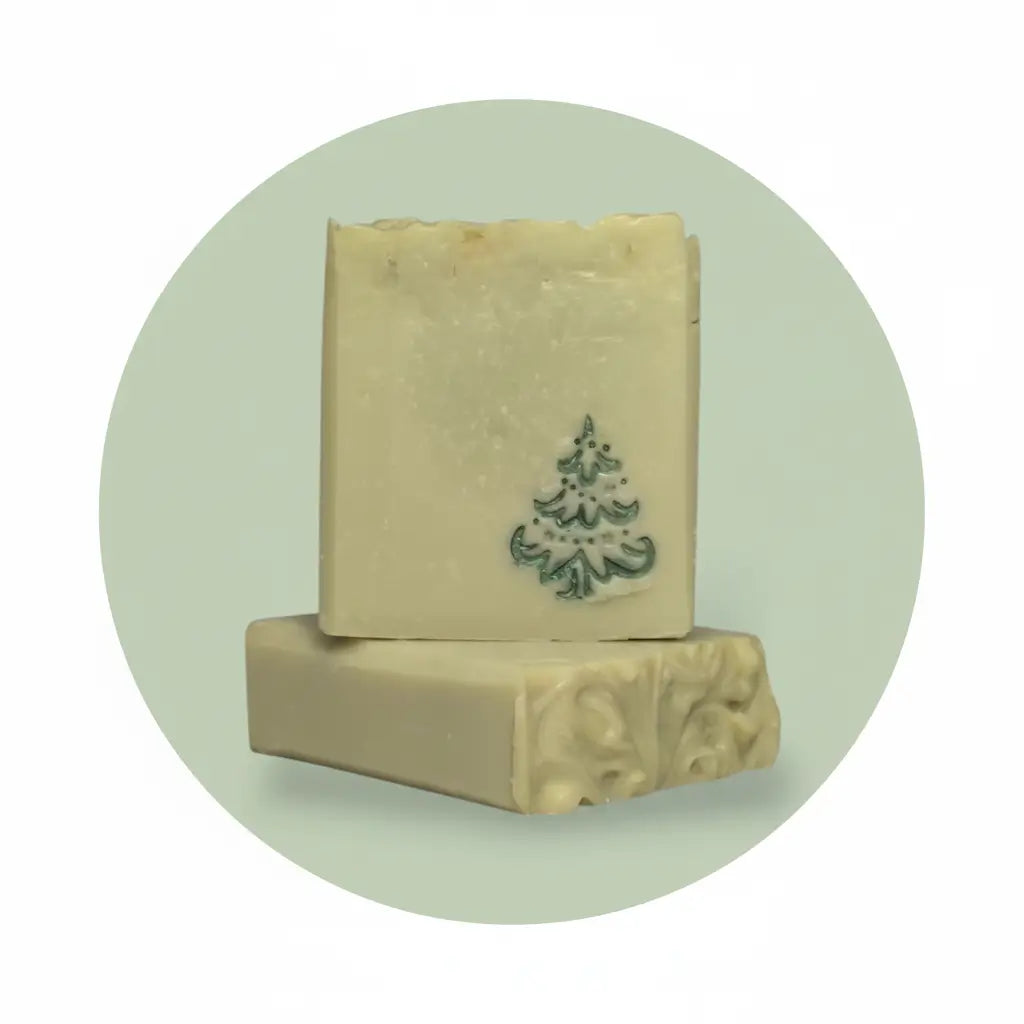Two rectangular bars of handmade soap stacked on a light mint green circular background. The top bar is cream-colored with a dark green hand-painted Christmas tree motif, and the bottom bar shows carved floral details on its side.