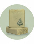 Two rectangular bars of handmade soap stacked on a light mint green circular background. The top bar is cream-colored with a dark green hand-painted Christmas tree motif, and the bottom bar shows carved floral details on its side.