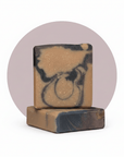 Two stacked rectangular handmade soap bars with a marbled swirl pattern of tan, brown, and dark charcoal, set against a white background with a muted mauve circular graphic behind them.