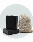 Two black rectangular bars of soap stacked on top of each other next to a beige jute drawstring pouch, set against a white background with a light blue-green circular graphic overlay.