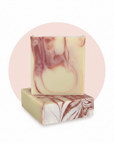 Two rectangular handmade soap bars stacked on a white background with a pale pink circular backdrop. The soap has a creamy off-white base with marbled dusty rose and mauve swirls.